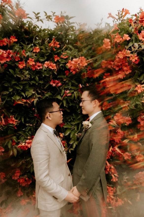 Two Asian people in suits stand facing each other, holding hands. The flowers and foreground are intentionally blurry, creating a vintage effect.