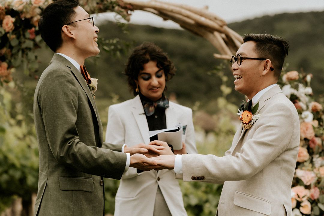 Two Asian grooms in glasses hold hands while exchanging vows. 