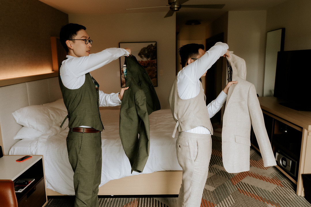 Two Asian grooms put on their suit jackets at the same time. One is wearing olive green and the other is wearing an oatmeal tan. They're in a hotel room.