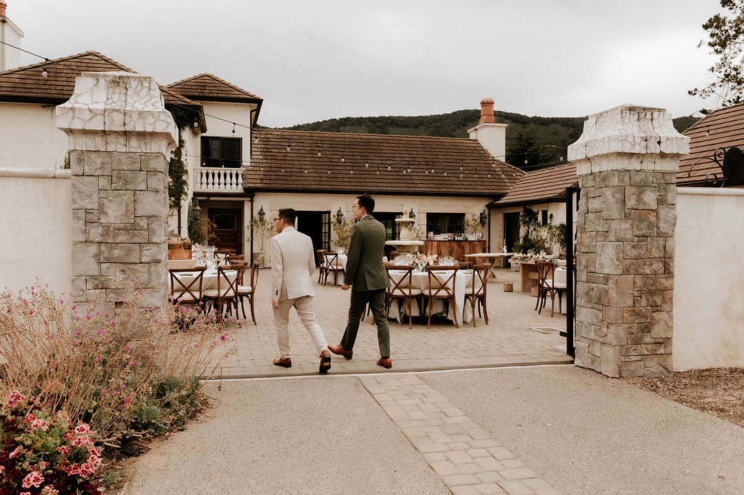 Grooms walk away from camera into the winery where they're getting married. One is in a cream suit and the other is wearing an olive green suit.