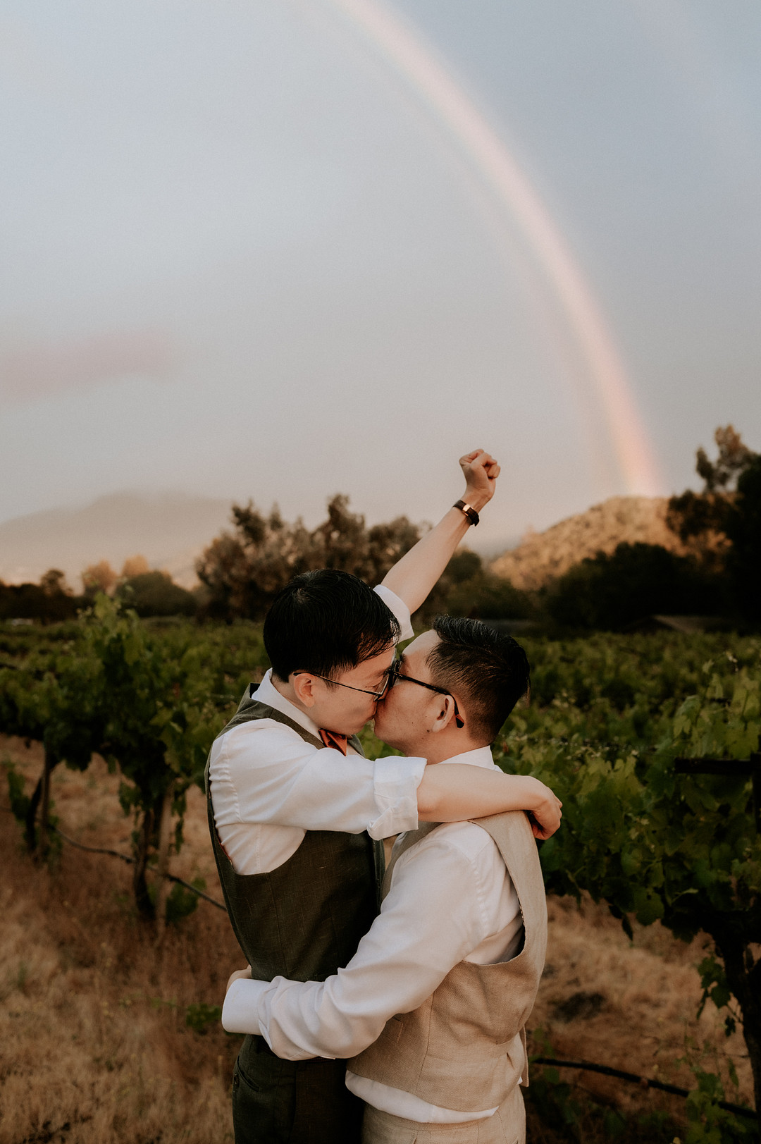 Asian grooms kiss while one holds his fist in the air. There's a rainbow behind them.