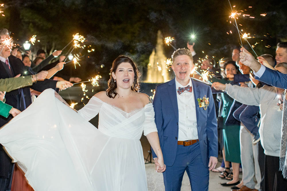 white nonbinary person in blue suit and white brunette queer bride walk through a row of wedding guests holding up sparklers for the sendoff