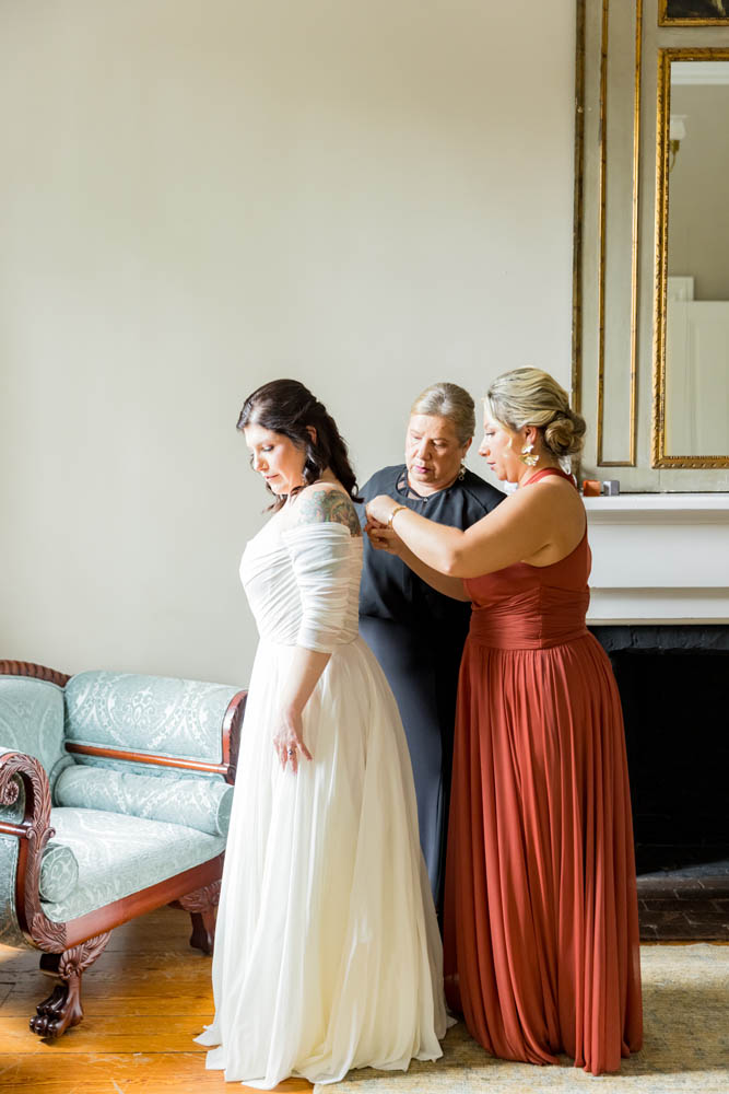 Three white women, one brunette in a white wedding gown, two blonde women helping her finish her wedding dress