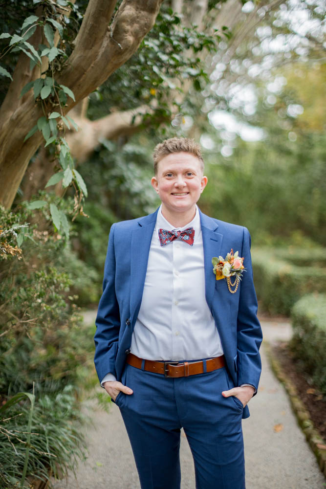 white nonbinary broom with short blonde hair standing outside, smiling. They are wearing a navy suit, a white shirt, bow ties, orange belt, wearing flower card in pocket as a boutonniere