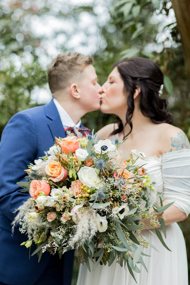 white couple kissing. one person has short blonde hair and is wearing a blue blazer. the other person has long dark brown hair and is wearing an off the shoulder white wedding gown and is holding a large bouquet of flowers with peach roses and anenomes