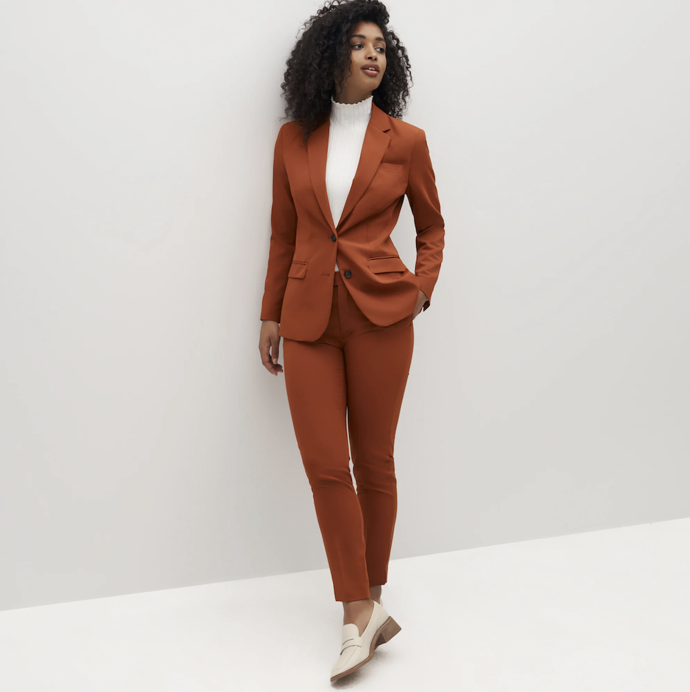 Black woman with natural hair wearing a burnt orange suit and a white mock turtleneck 