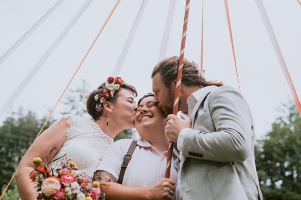 throuple wedding for three white people. Two white women with short brown hair with a white man with blonde hair in a french braid all smile and kiss while holding onto ribbons