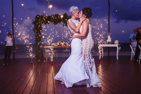 Two white lesbian women, one with cropped platinum hair and the other with long brunette hair in an up do, kiss while holding each other with cold fireworks in the background.