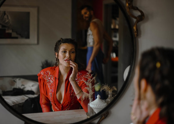 A white person in a red fitted top sits and looks at themselves in a round mirror. They are touching their lips and there is another person in the reflection behind them