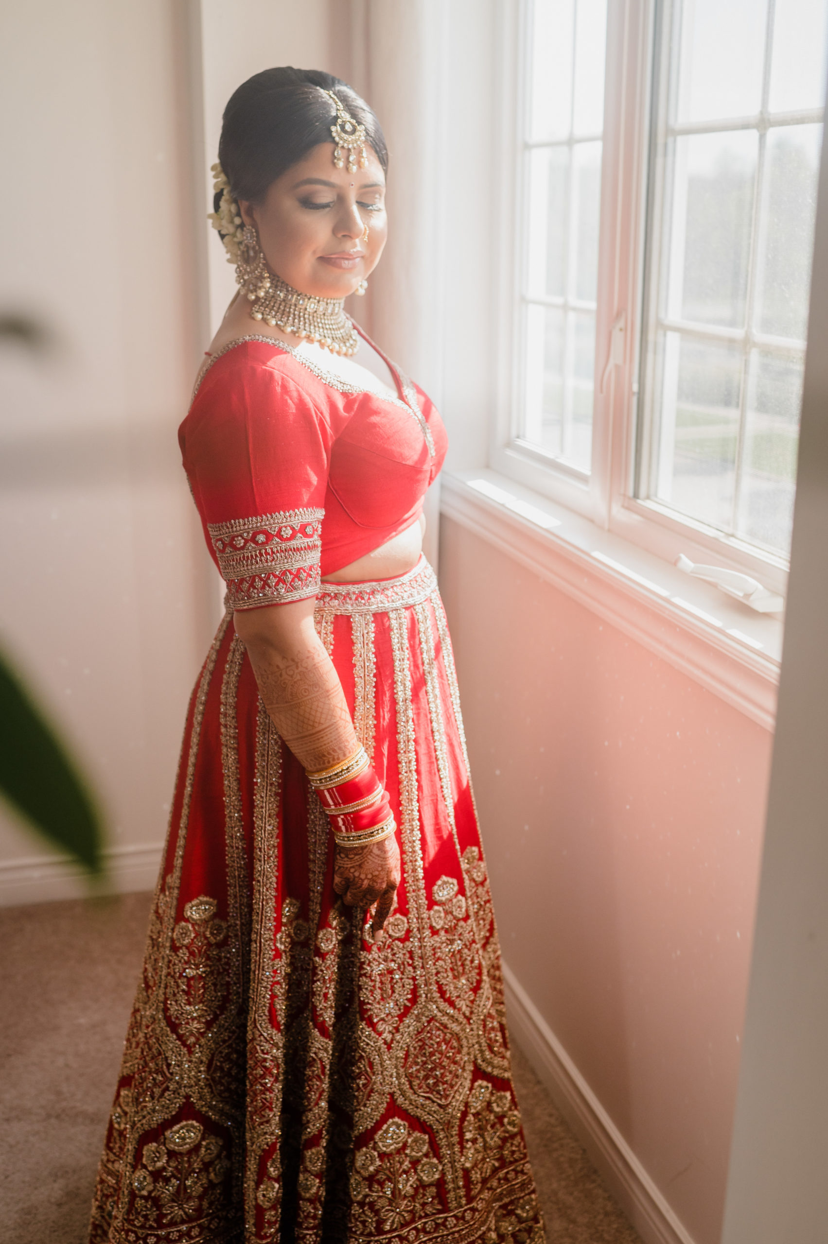 A South Asian person in a red lehenga stands by a window with their eyes closed, showing off their traditional gold adornments.