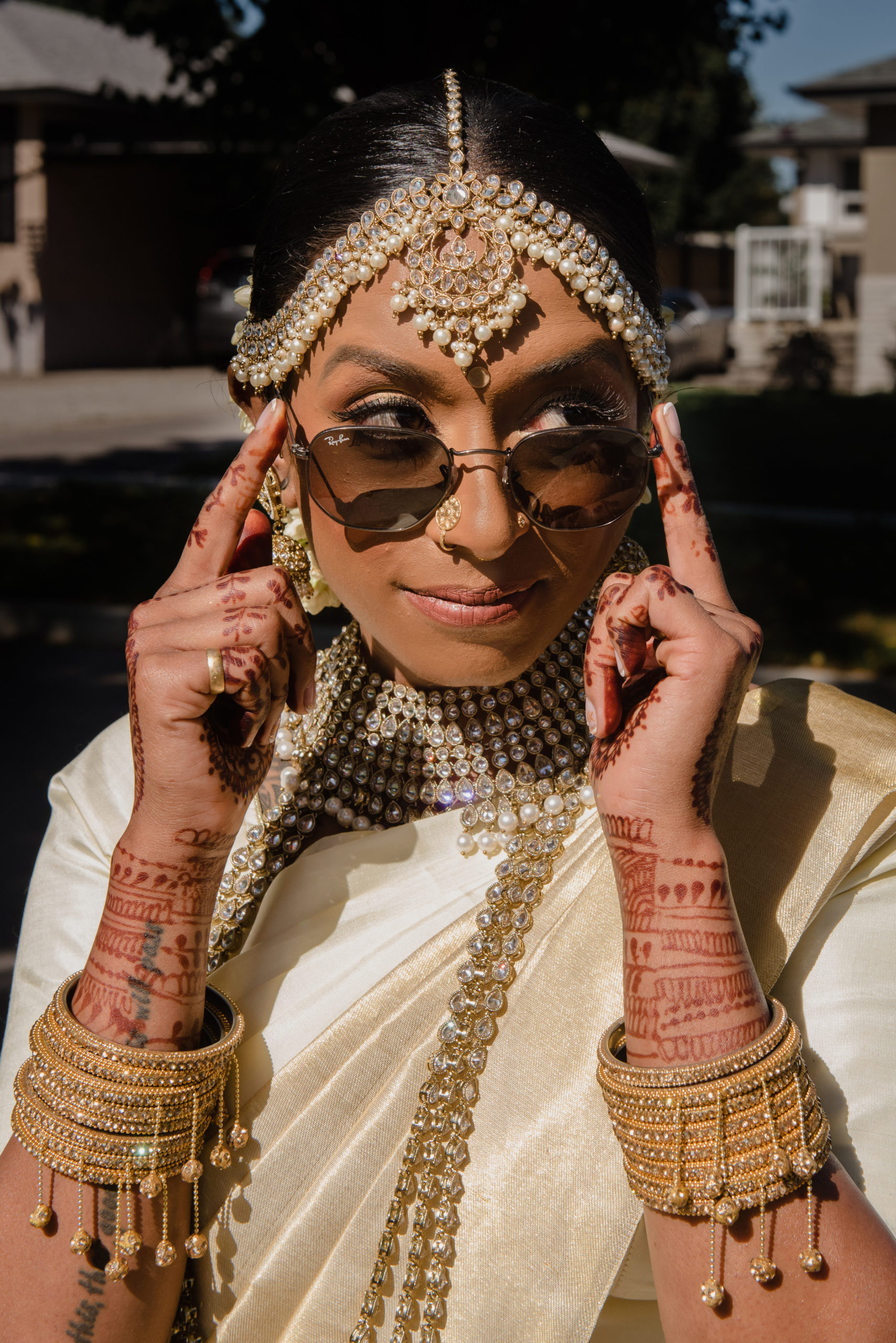 A South Asian person with wedding henna and jewelry holds their sunglasses and looks to the left.
