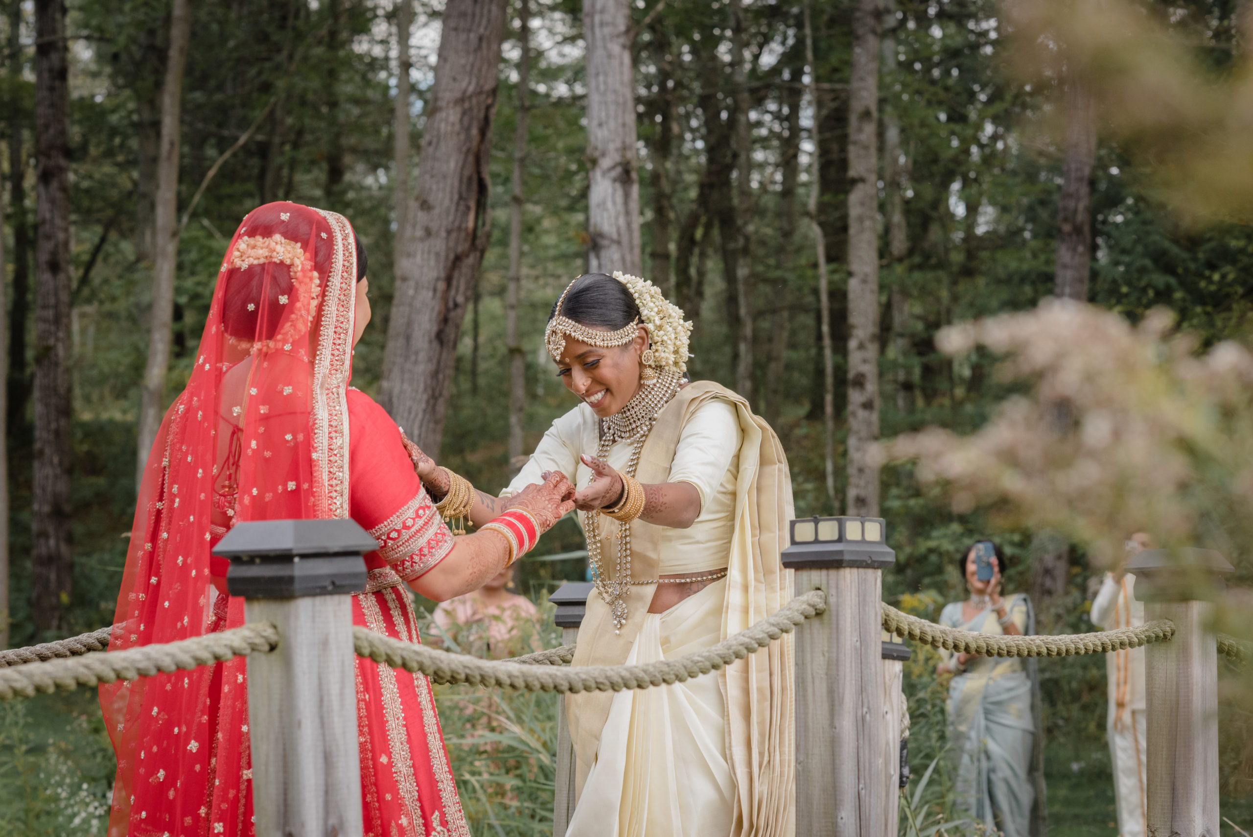A pair of South Asian marriers stand on a footbridge, admiring each others' wedding attire. The one on the left is wearing red, and the one on the right is wearing tan and gold.