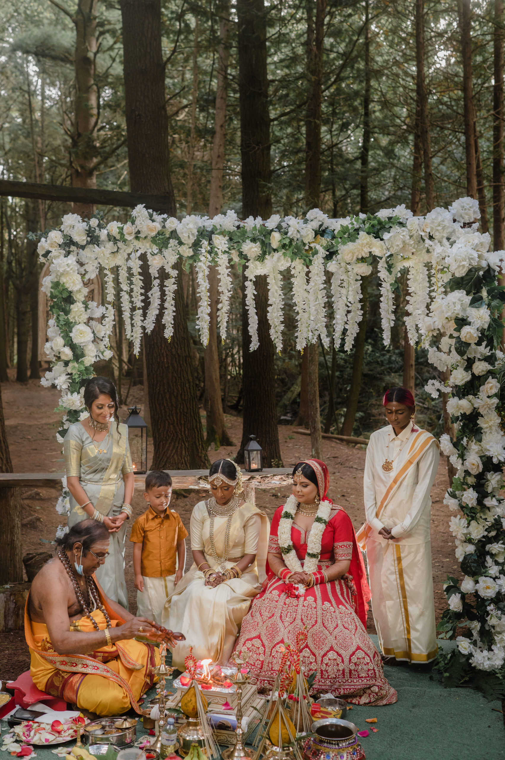 A pair of South Asian marriers kneel under a white floral arch as they watch their officiant perform a traditional ritual.