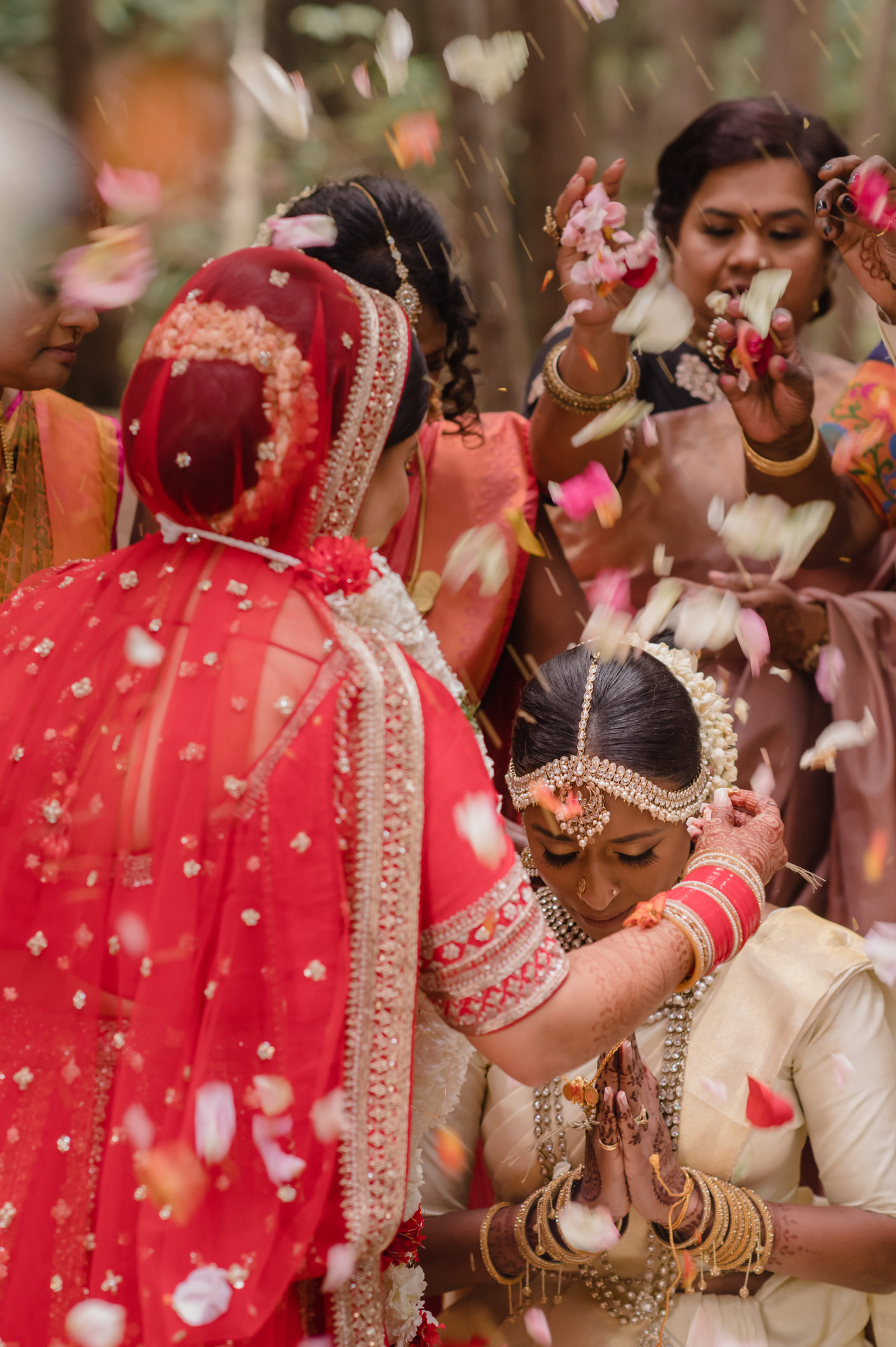 A South Asian person kneels in front of their spouse as family throws rose petals on both of them. The person in the front stands and is wearing a red lehenga.