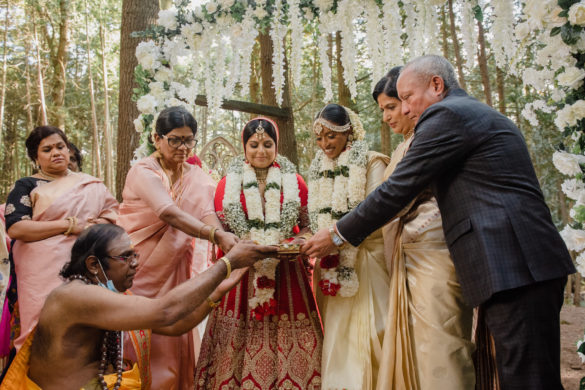 Two South Asian people stand under an arch of white flowers. They have loved ones surrounding them, all reaching to the couple's hands. The two newlyweds are wearing white floral necklaces.