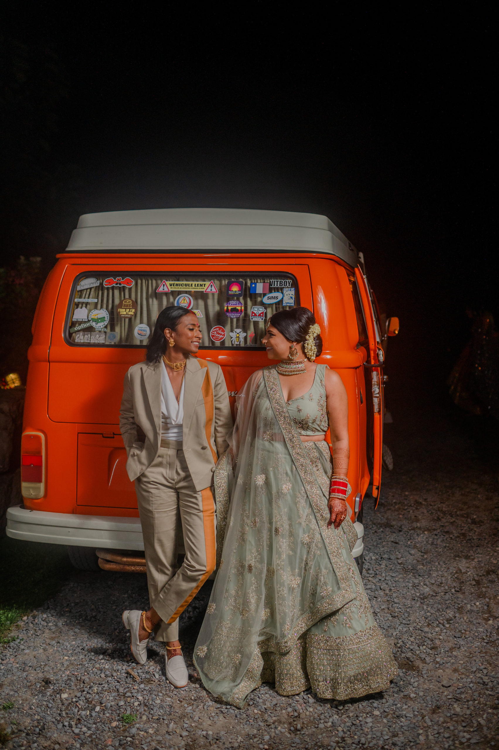 Two South Asian spouses stand in front of an orange camper van. The one on the left is wearing a tan suit with orange stripes, and the other wears a tan lehenga.