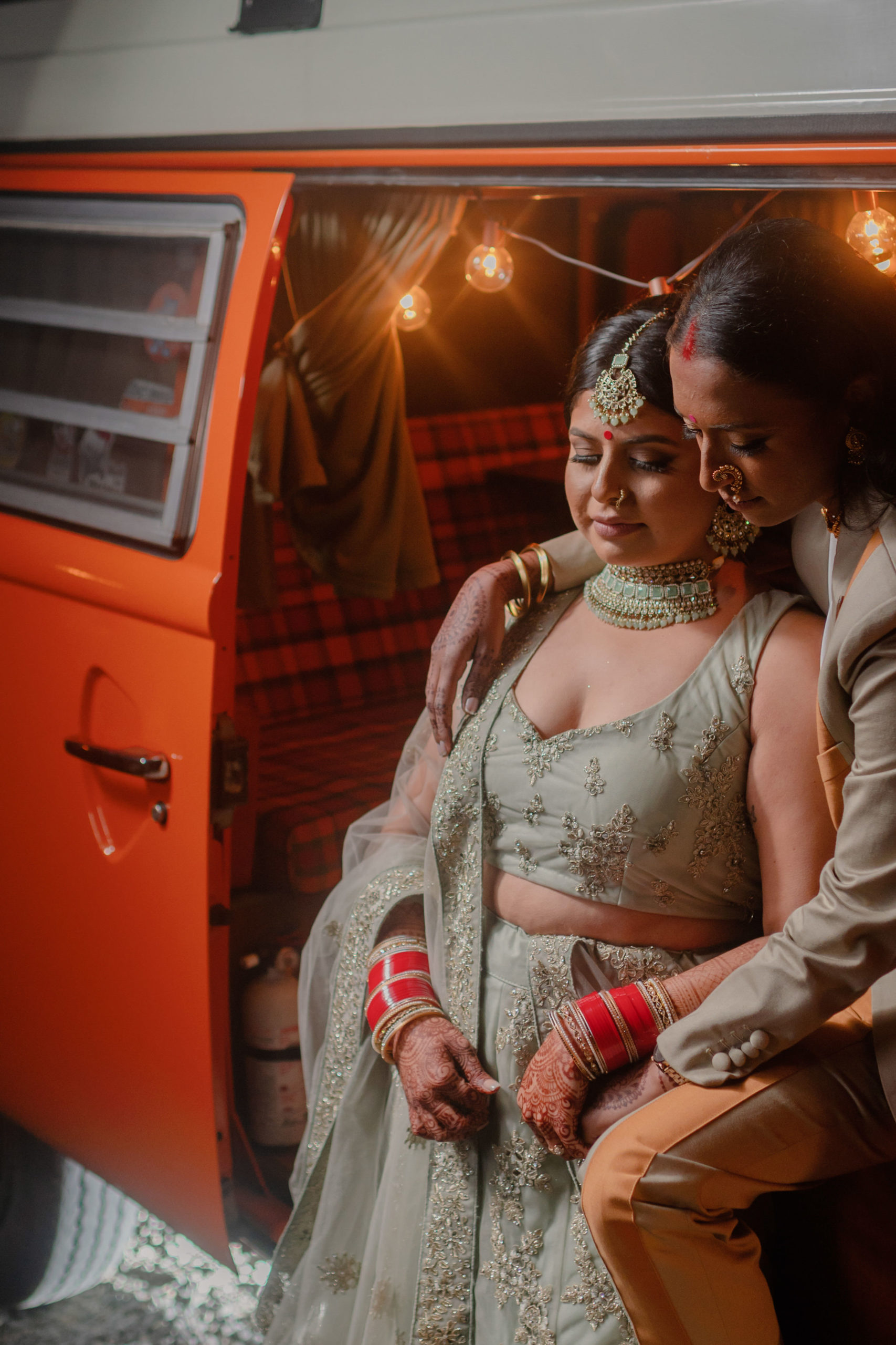 Two South Asian spouses sit in the door of an orange camper van. There are string lights behind them. Both are looking down at the henna decorations on their hands.