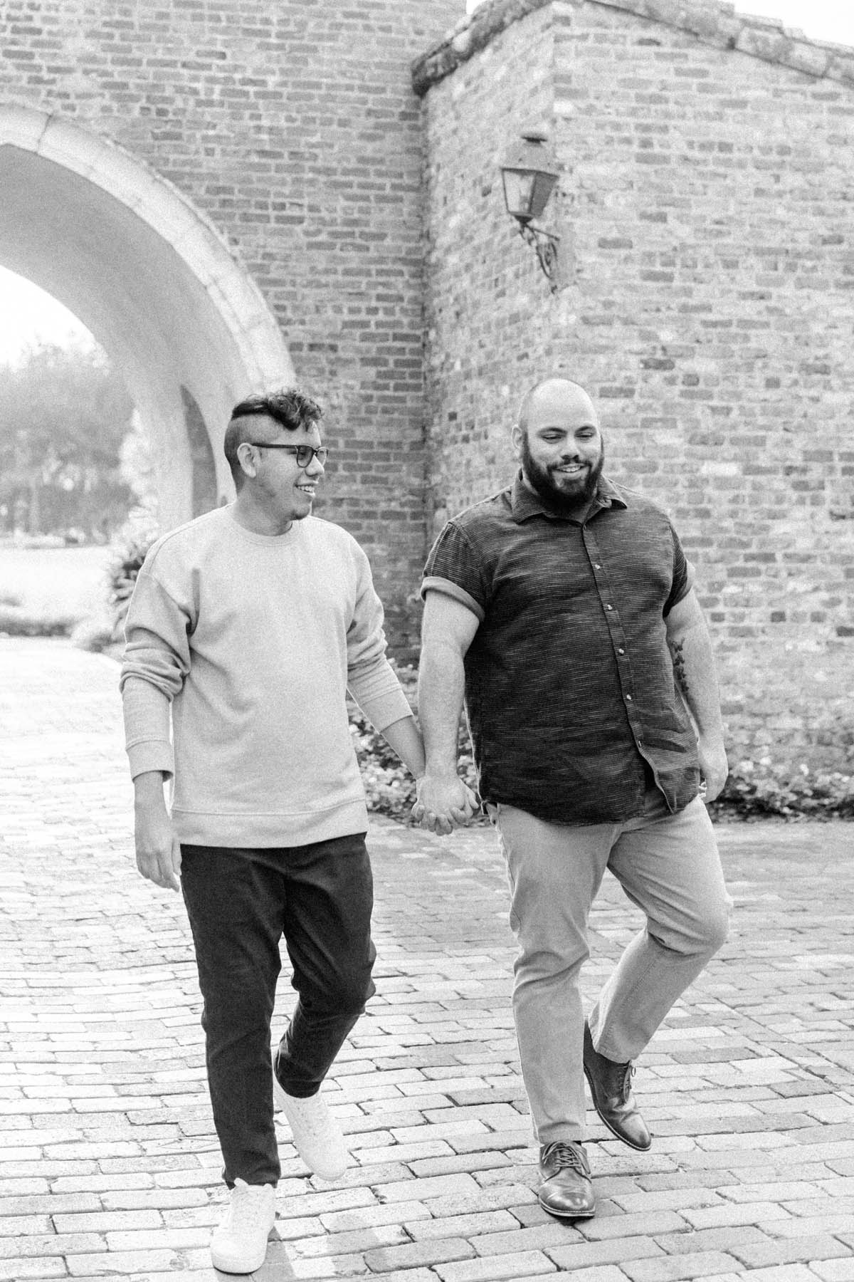 A black-and-white photo of two men holding hands in front of a brick building. 