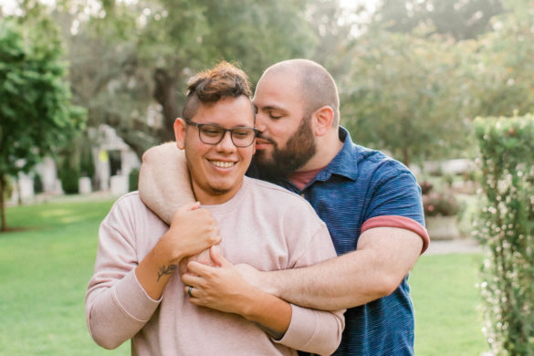 Two men stand in a green open space. One is Latinx and wears glasses. Behind him, hugging him is a man with facial hair wearing a blue shirt.