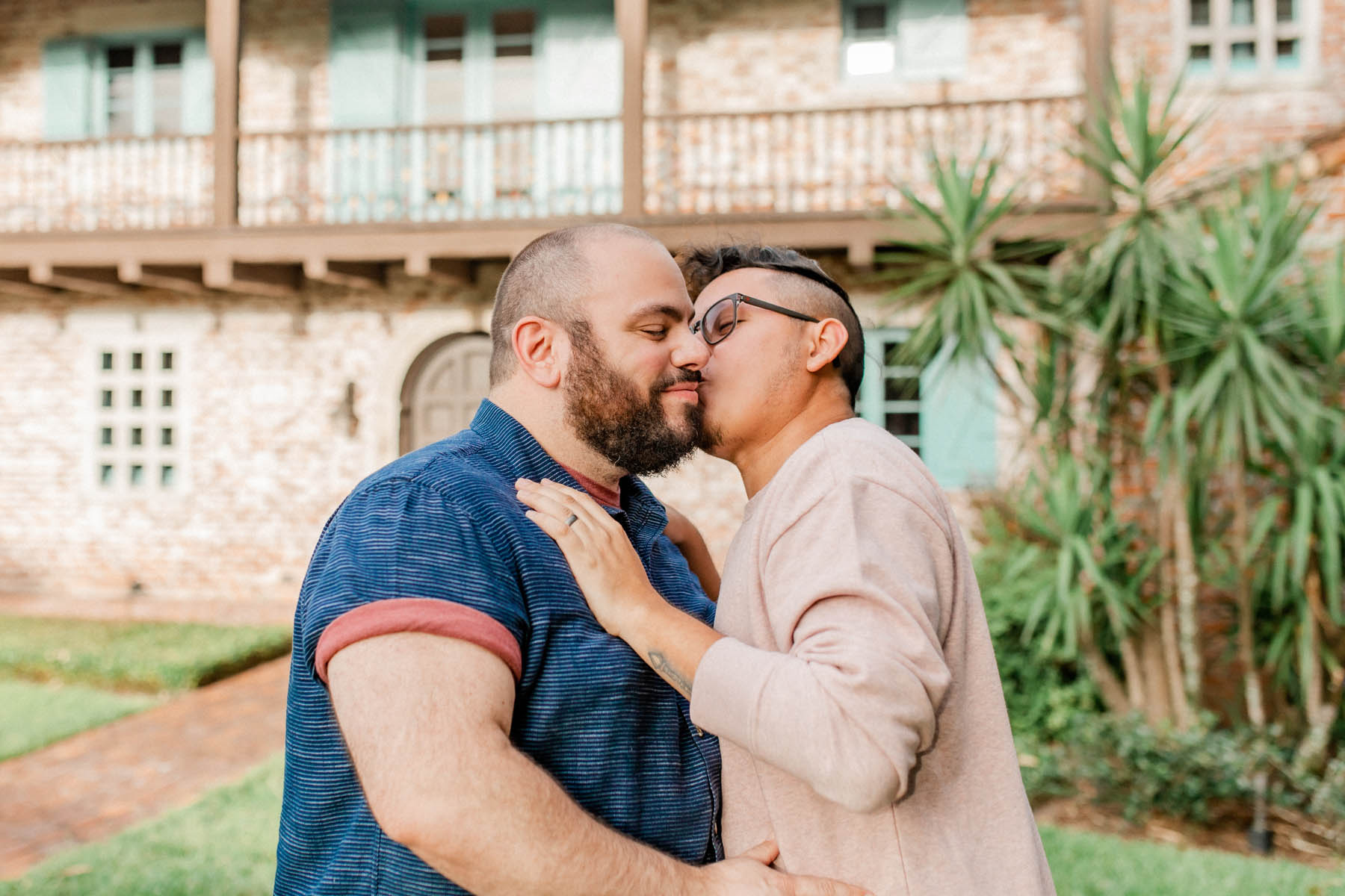 Two engaged men stand in front of a stone building. One kisses the other on the cheek. 