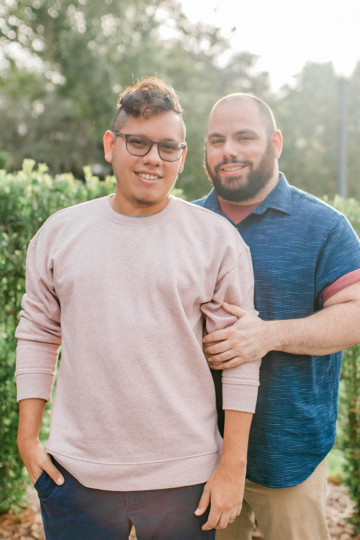 Two engaged men stand in an open garden. One is wearing a pink sweater, the other wearing a blue shirt. They are smiling directly at the camera. 