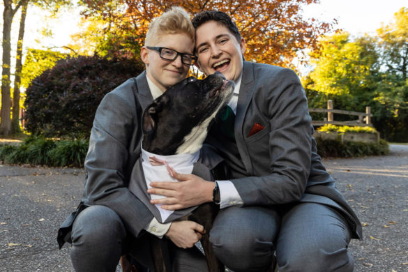 Two white people in suits pose with their dog, smiling at the camera. The dog is looking up and licking one of their faces.