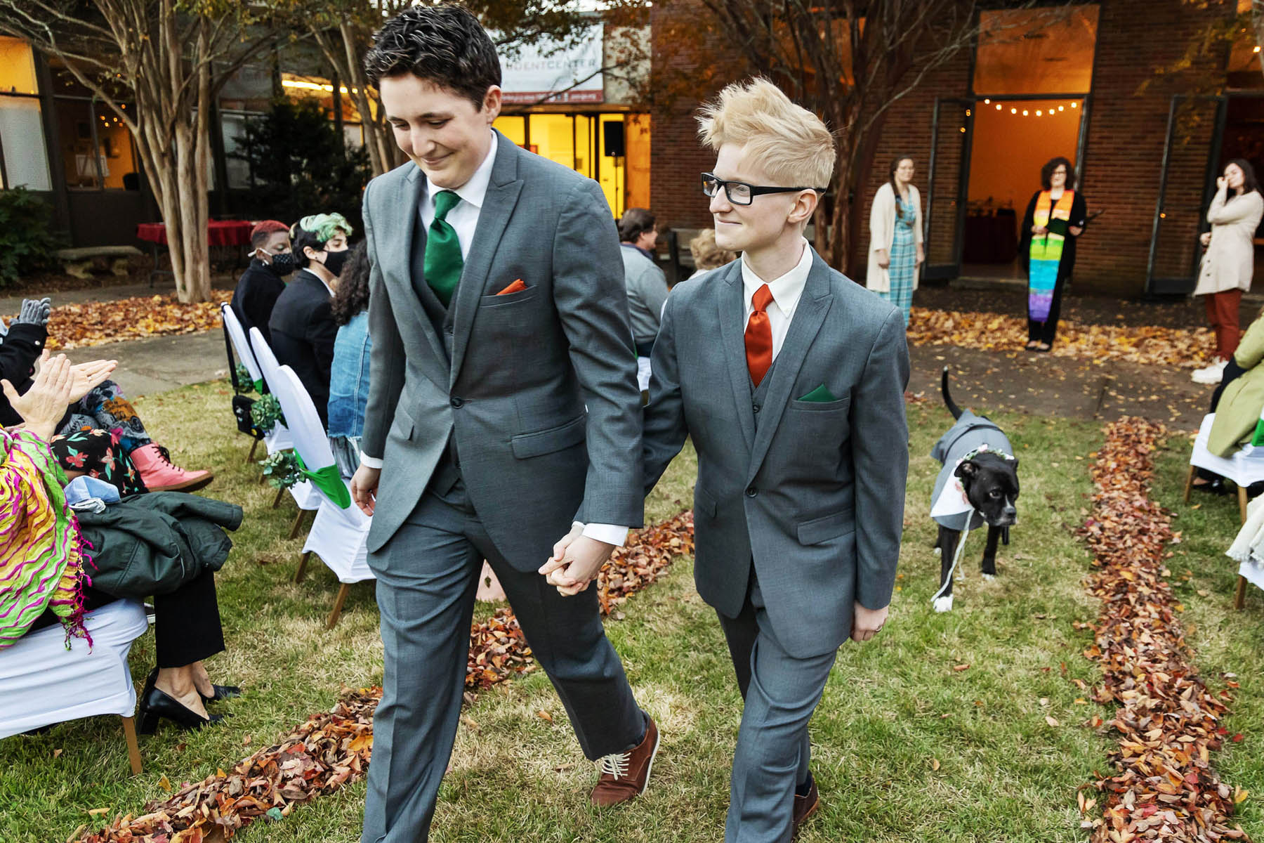Two white people walk down an aisle lined with fall leaves. Their dog, dressed in a little tuxedo shirt, follows them. 