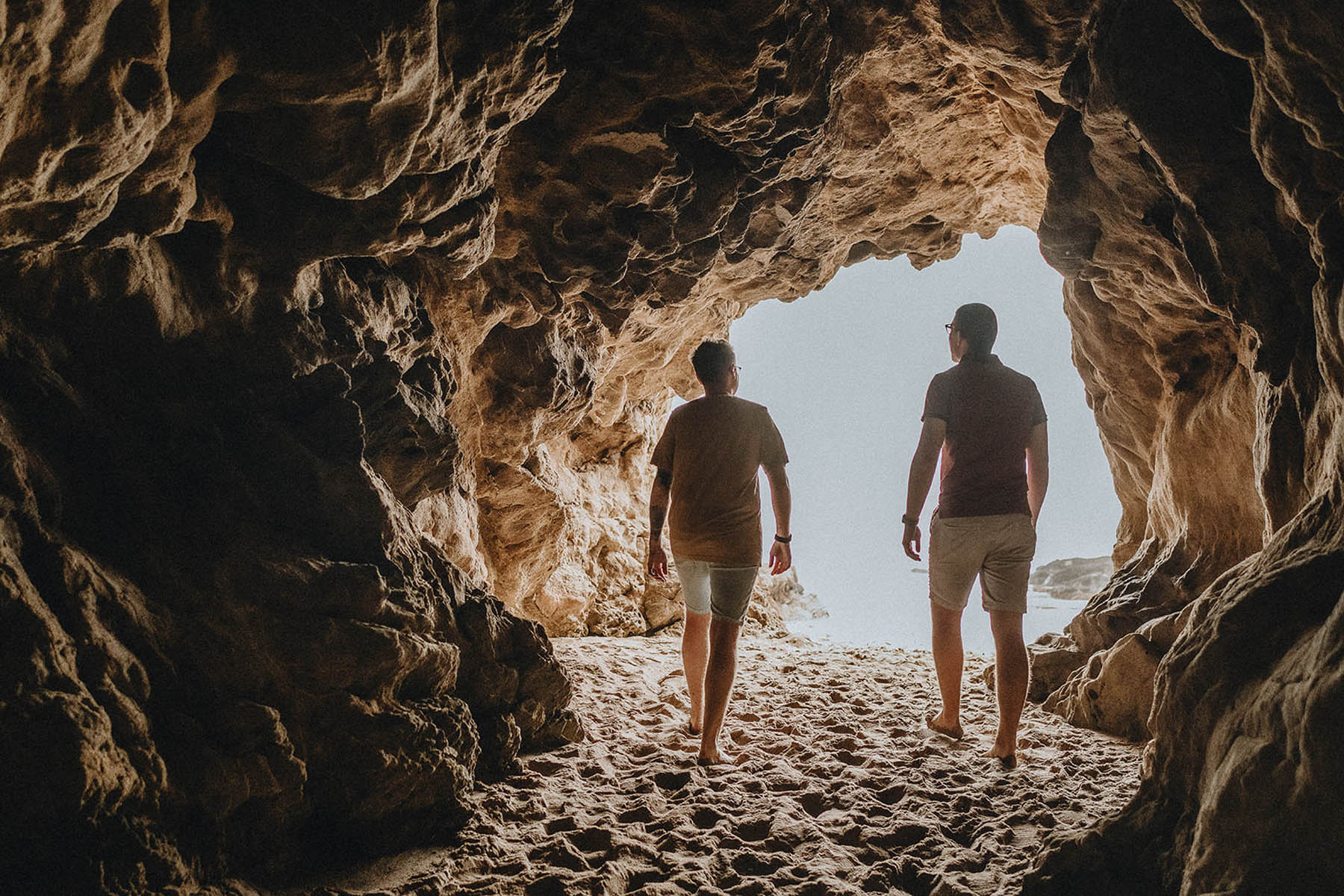 Two people walk through a cavernous rock cave looking out on an ocean.
