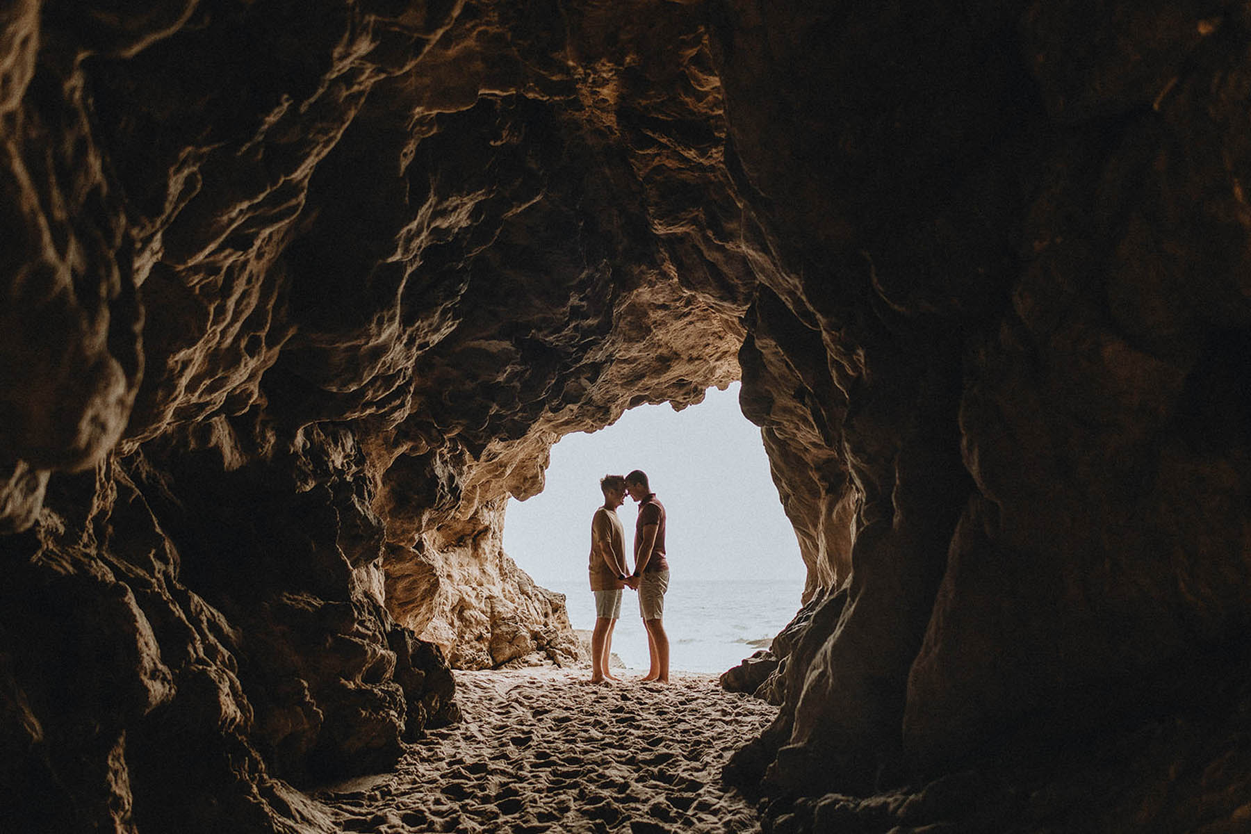 Two people stand, foreheads touching, holding hands in a large rock cave.