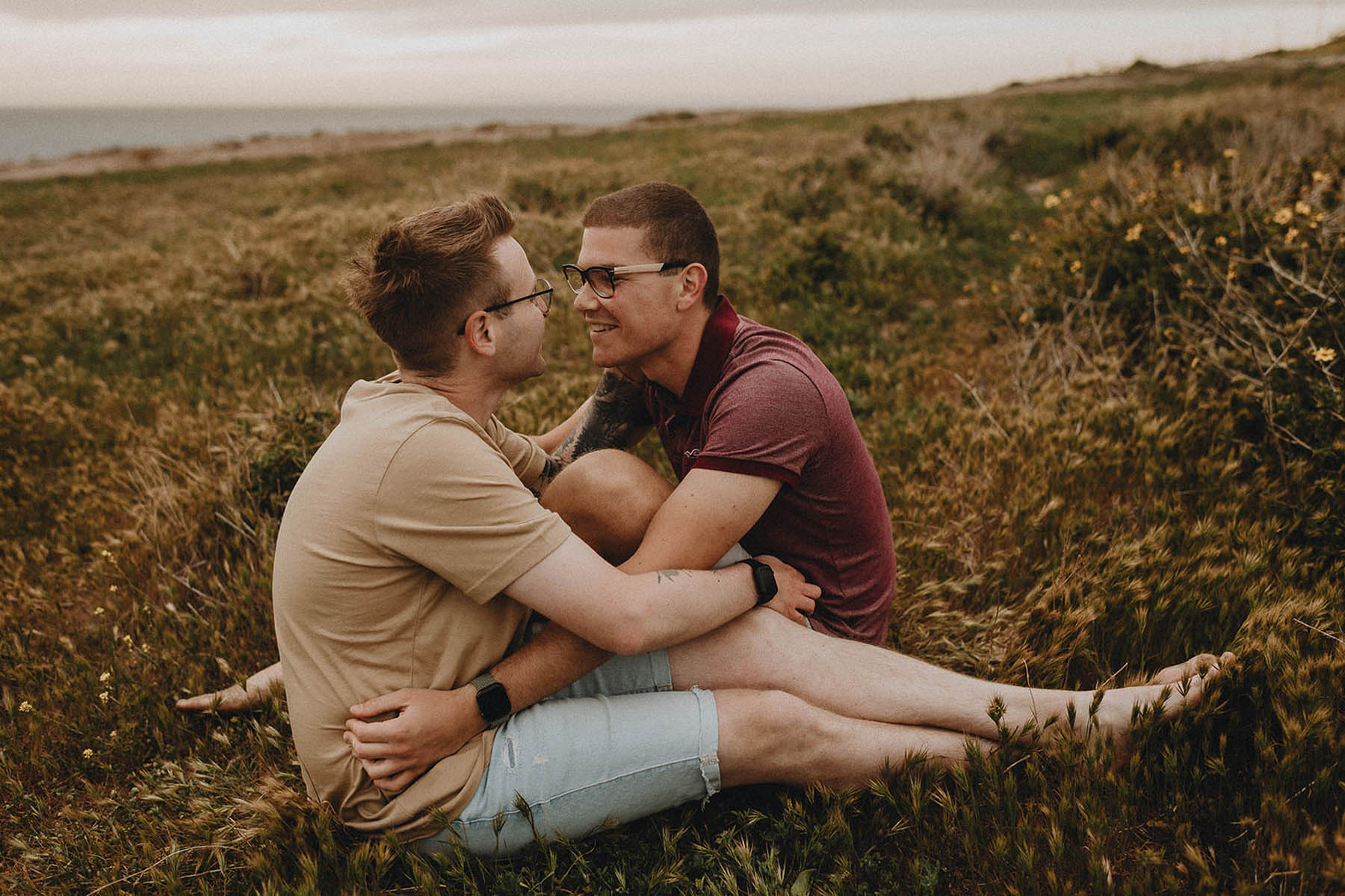 Two men sit facing each other in a meadow of wildflowers and grass.