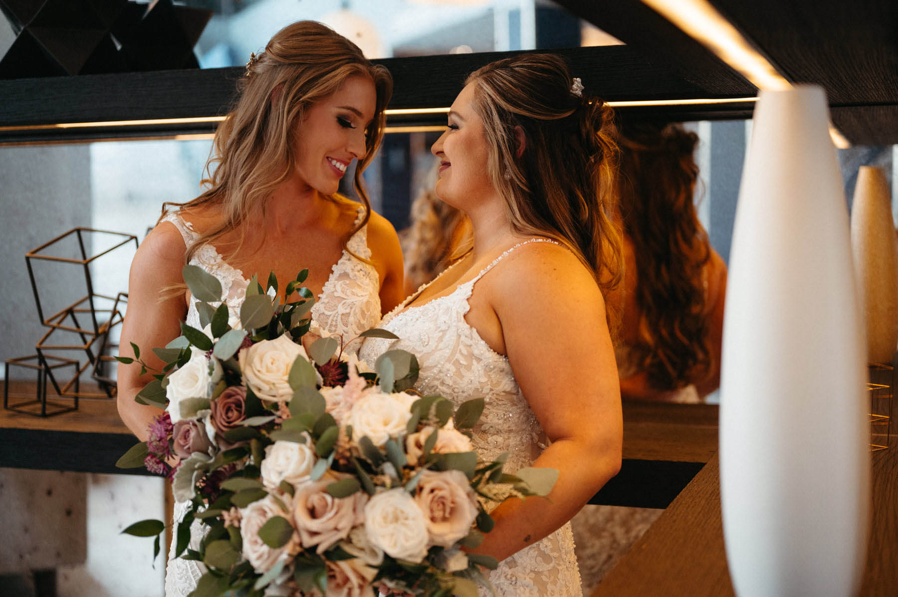 The brides stand in front of a mirror with their bouquets, smiling at each other.