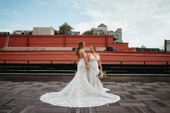Two white brides with long lace gowns stand on a NOLA rooftop with a red building in the background.