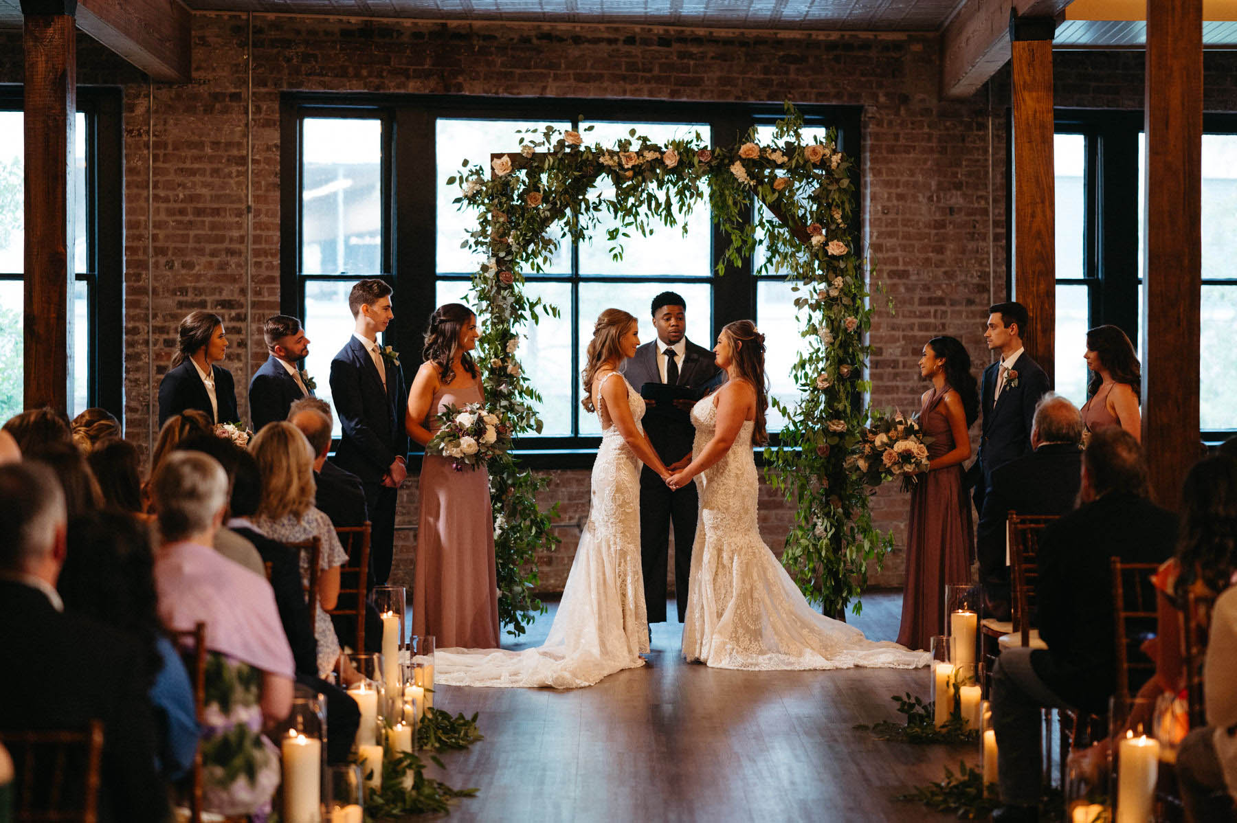 The brides stand under an arbor of greenery and hold hands as they exchange vows.
