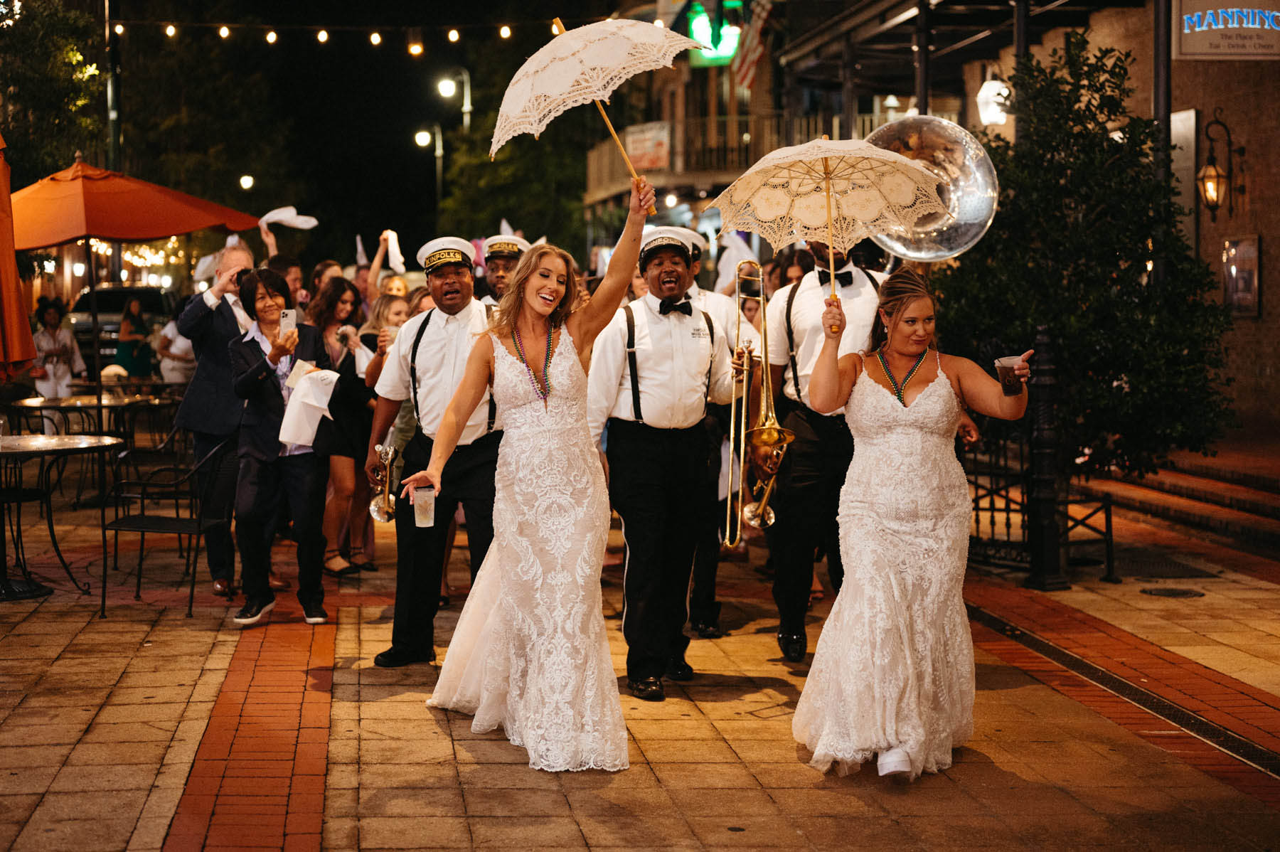 The brides lead a festive second line. They are holding white umbrellas and followed bu a jazz band.