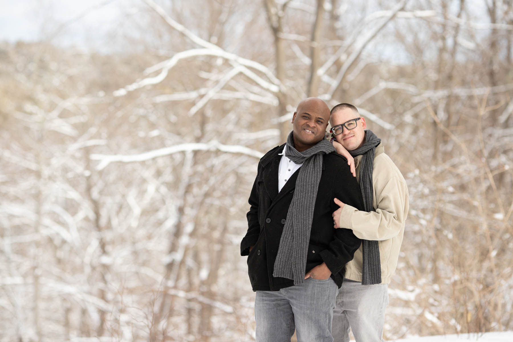 Two people stand in front of snowy trees, smiling at the camera. The person on the right holds the arm of the person on the left.
