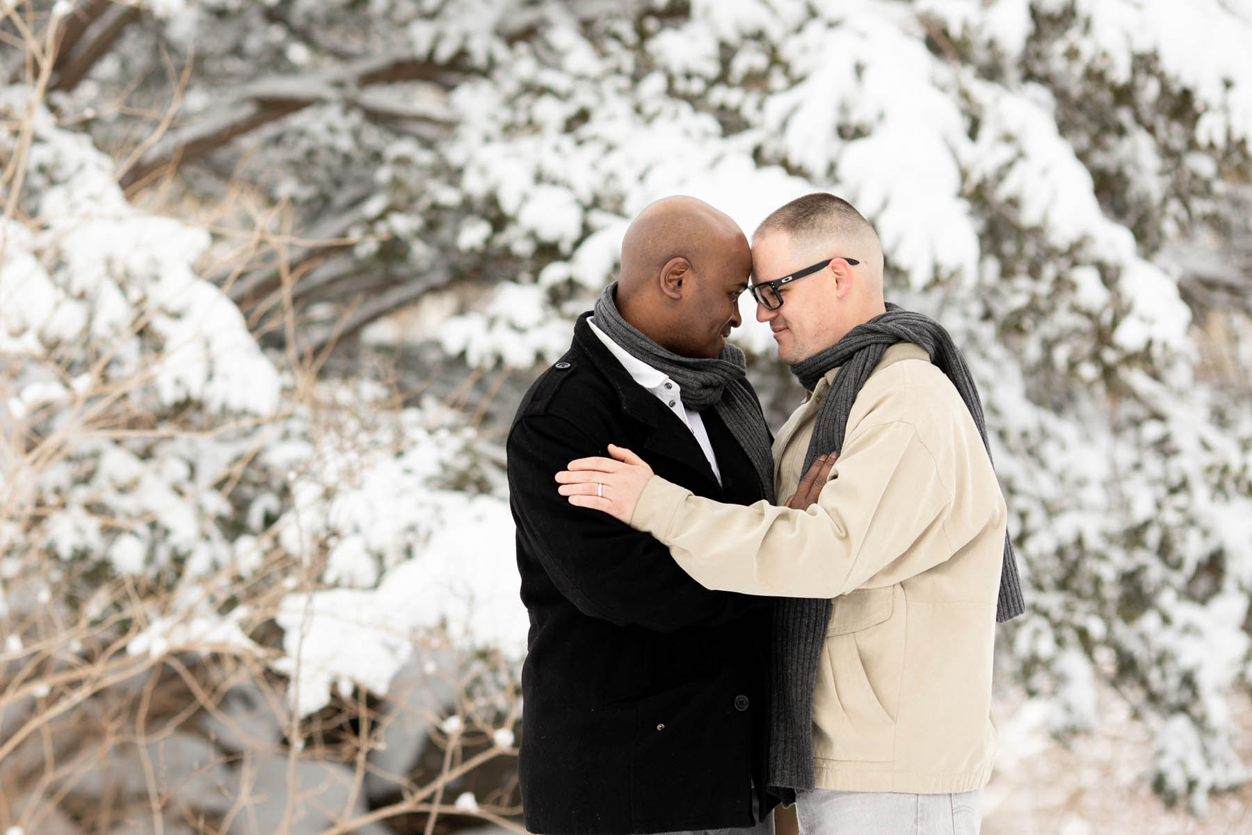 Two men stand in front of a snowy background. They are both wearing matching scarves and gazing into each others' eyes. 