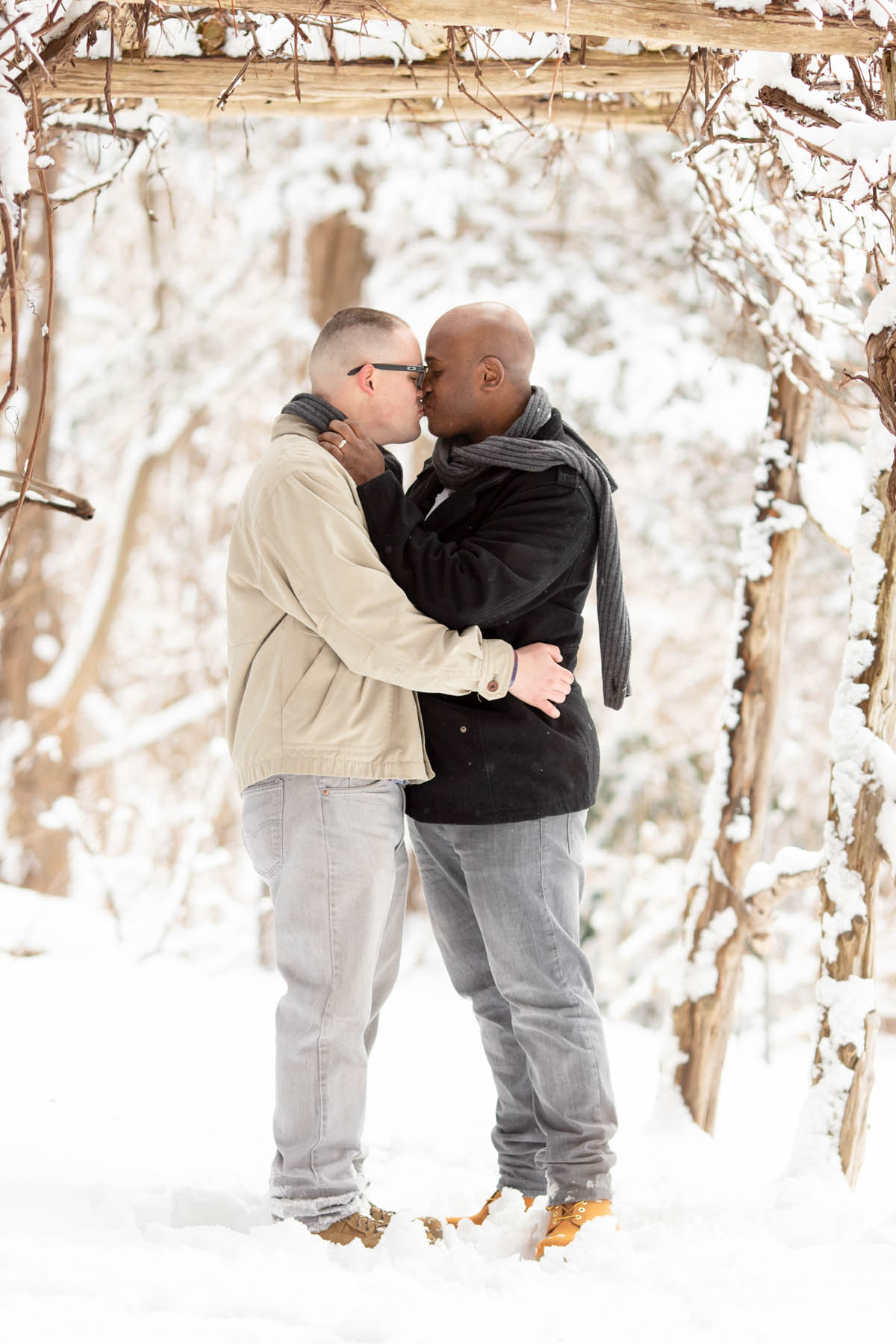 An engaged couple stands under a wooden arch covered in snow. They are kissing and facing each other.
