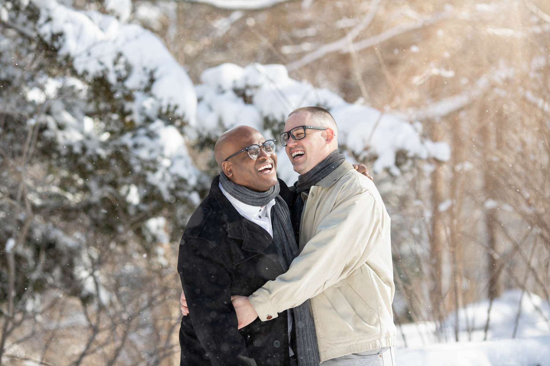 A black man and white man stand, embracing. They are both laughing as snow falls in front of them.
