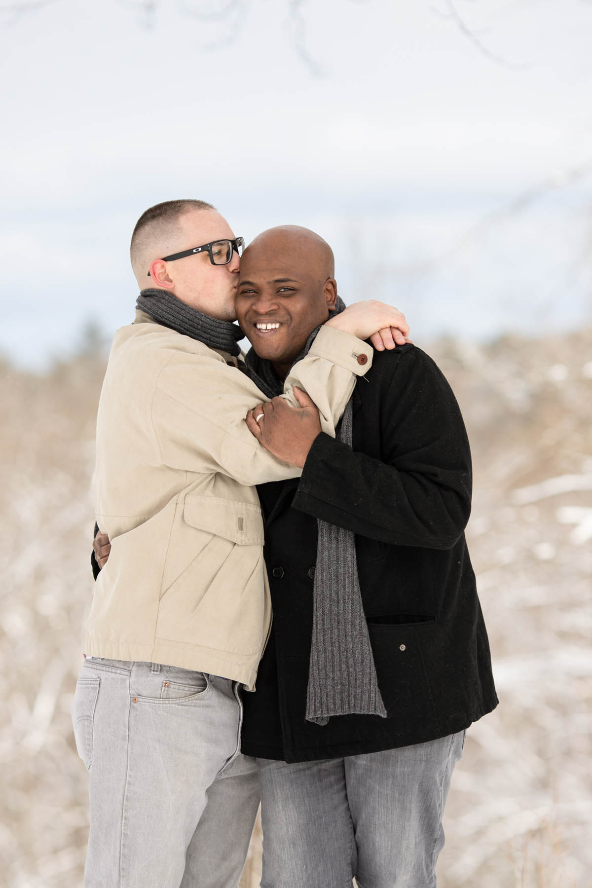 A man in a tan jacket is kissing the head of a man in a black jacket. The man on the right is smiling at the camera.
