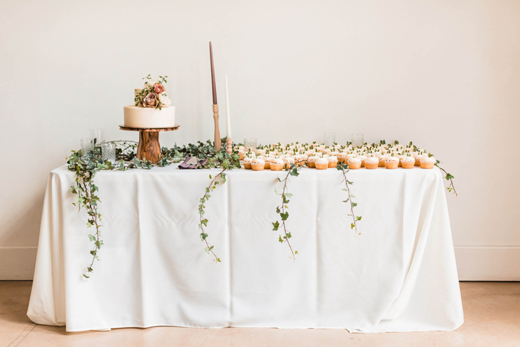 A rectangular table with a white tablecloth holds a small cake and cupcakes. There are two candles and many strands of greenery around the table.