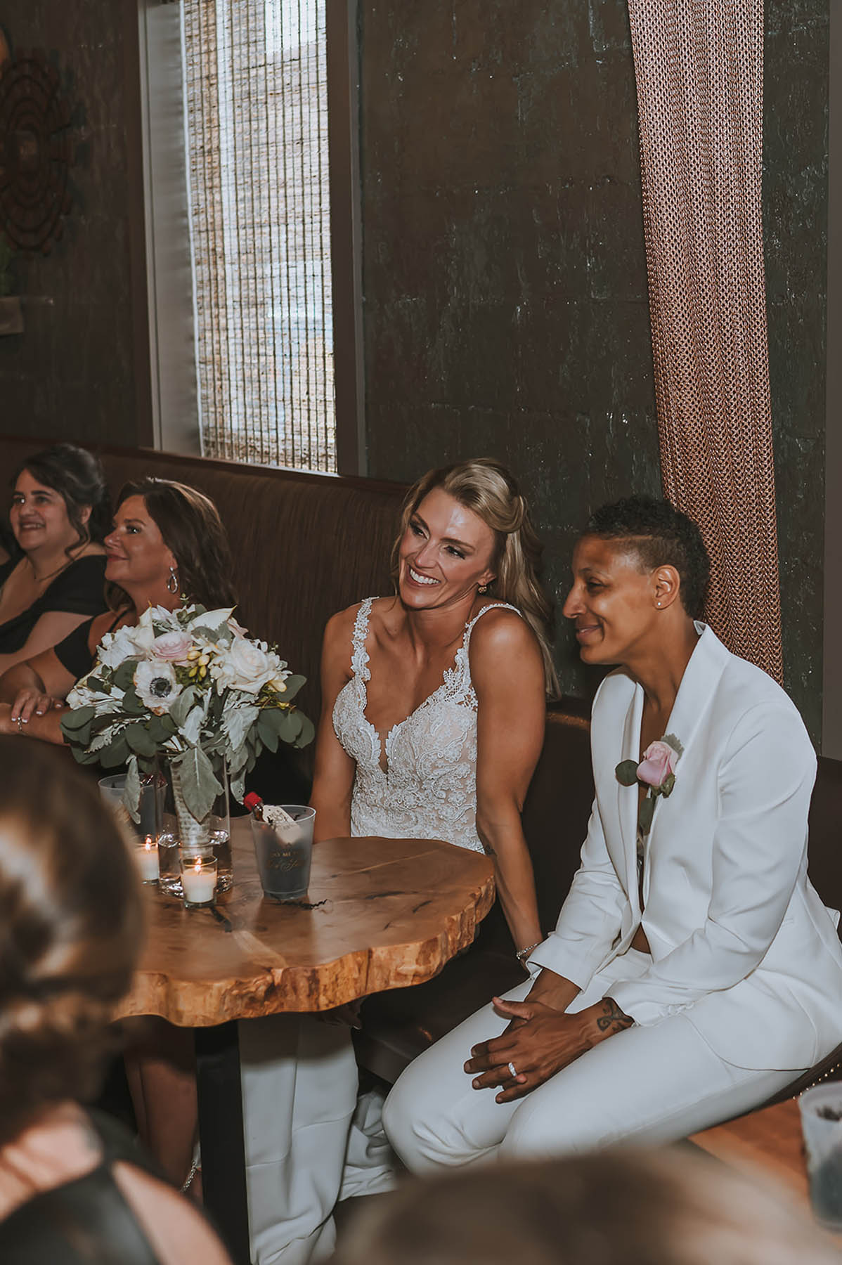 The newlyweds sit ad a small bistro table and smile as they listen to a speech.
