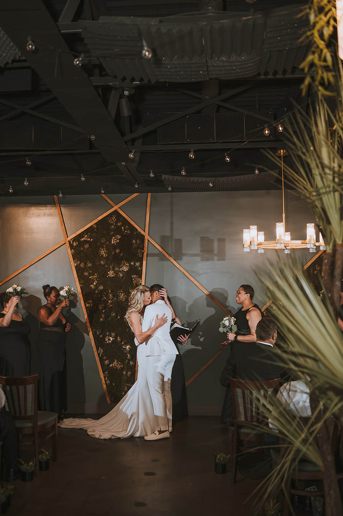The newlyweds share a kiss in front of a geometric backdrop as their wedding party cheers.