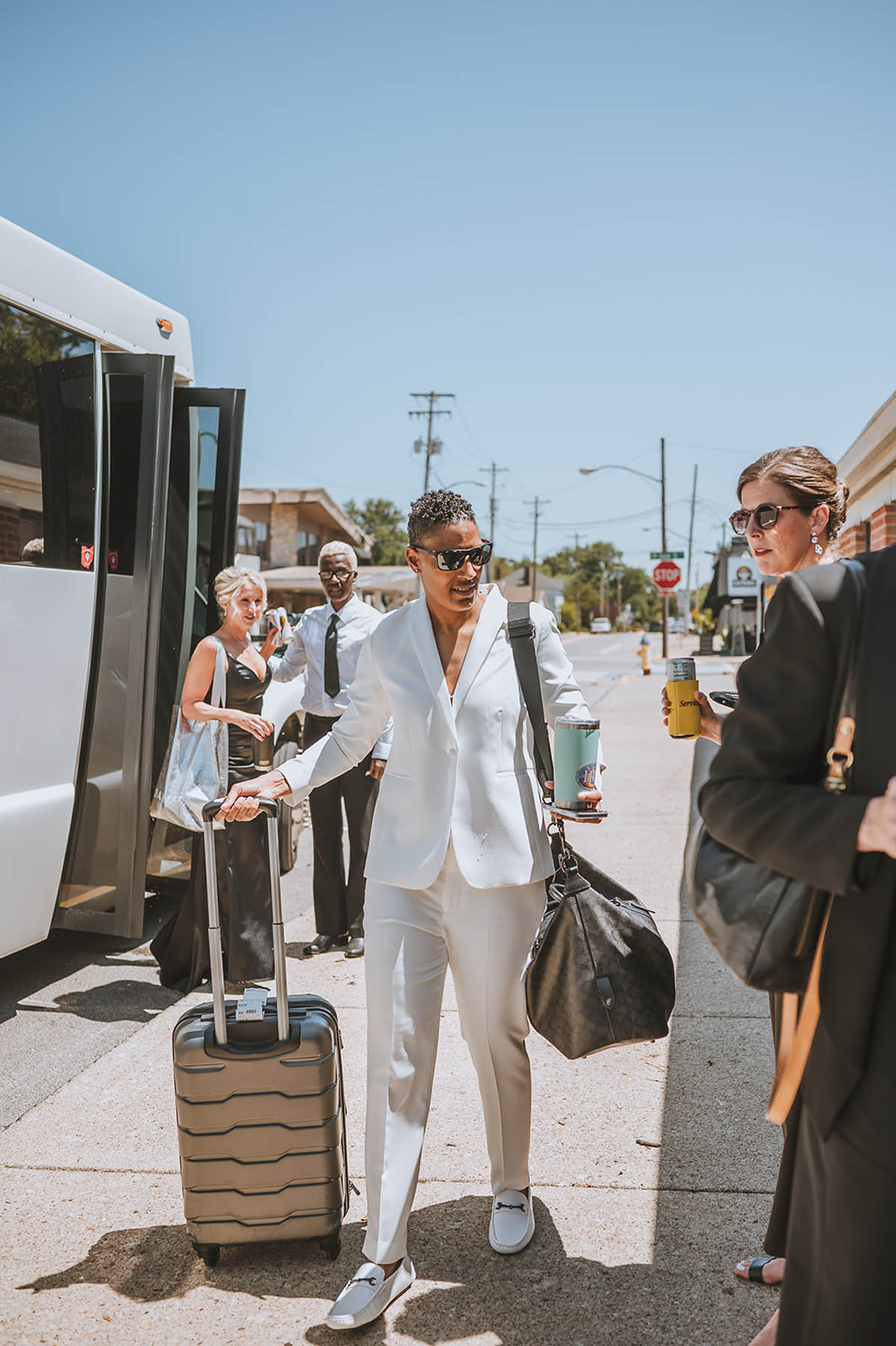 One of the marriers arrives off a party bus, wearing sunglasses and a sleek white suit and carrying luggage.