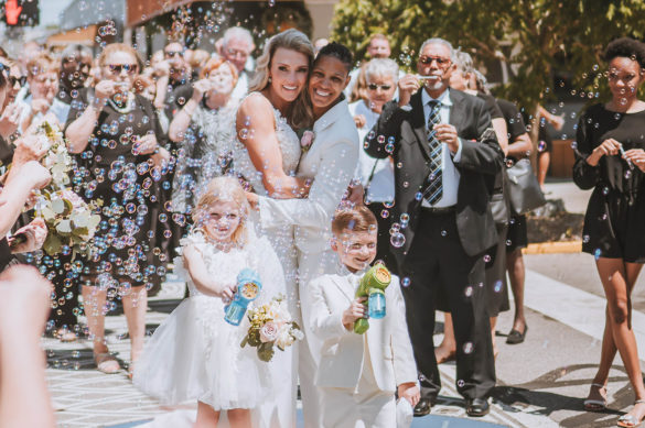 Two brides, one white and blonde and one black and with short dark hair, stand embracing with their celebrants around them. They are smiling, and there are bubbles surrounding them.