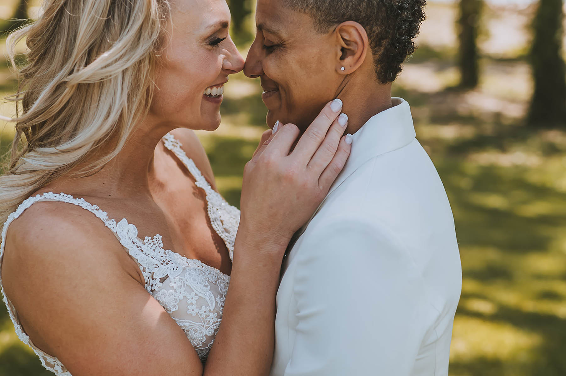 An interracial couple embraces, noses touching, in their white wedding attire.