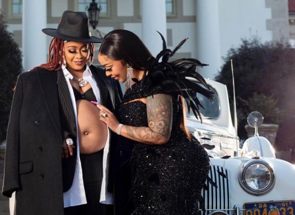 Two black women, dressed in black, stand in front of a stately mansion and cream-colored car. The one on the left is Da Brat, and is wearing a hat and unbuttoned top that shows off her baby bump.