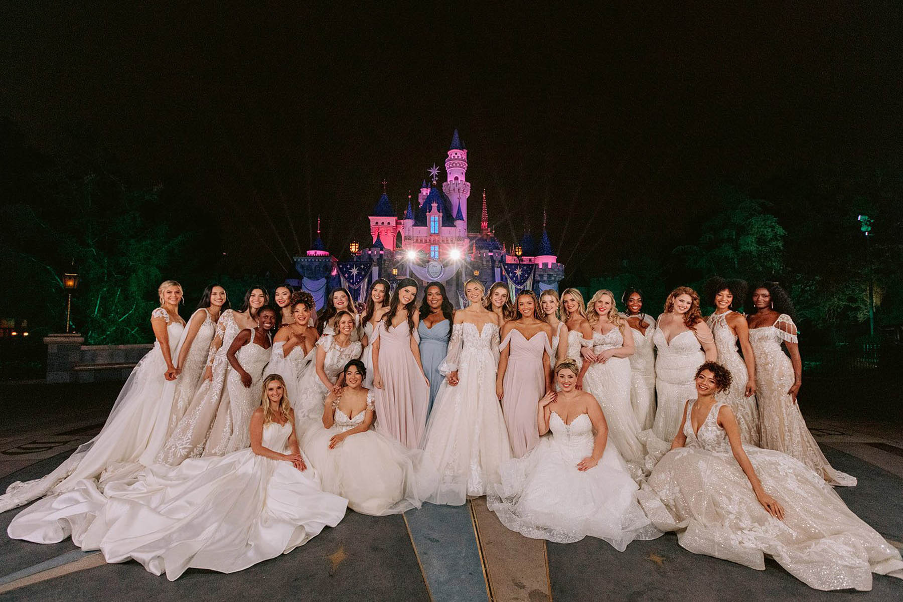 A large group of people in princess-inspired gowns poses in front of Sleeping Beauty's castle.