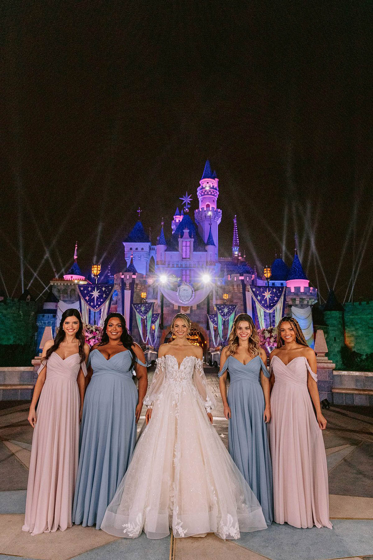 A group of people in a white gown and pink and blue wedding party dresses stand in front of Sleeping Beauty's castle.