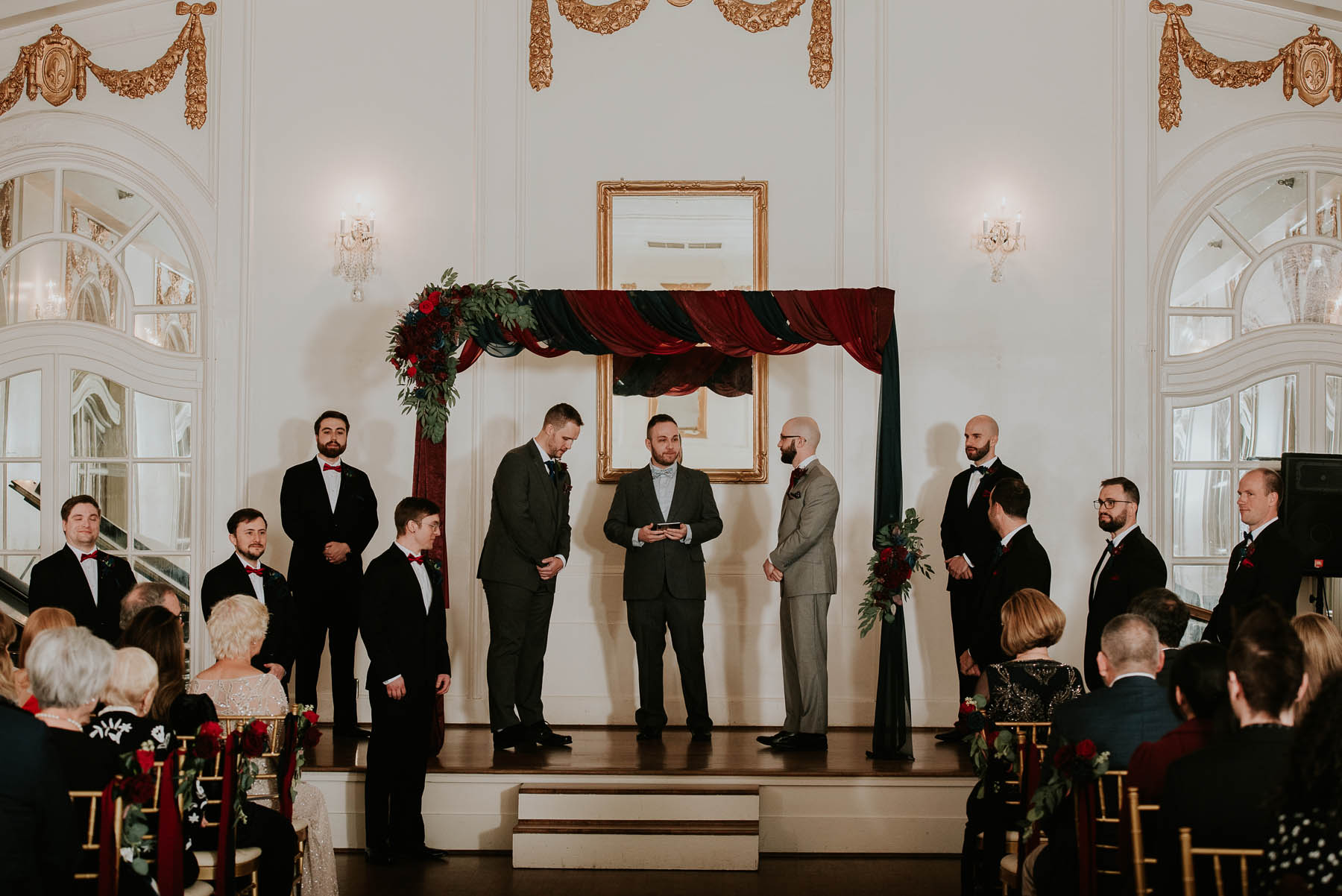 Two white men stand under a burgundy draped arbor and exchange vows.