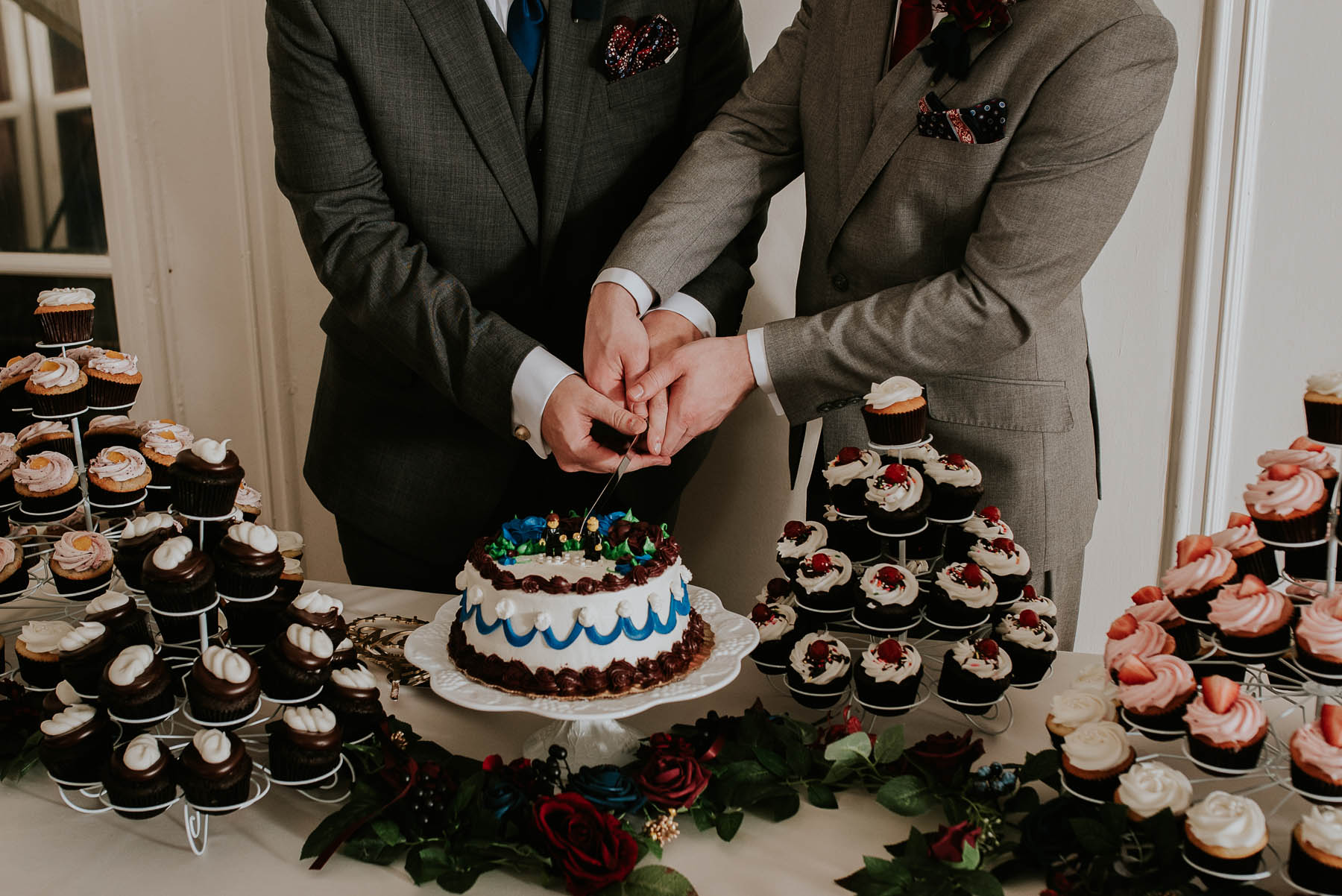 The grooms cut into a single tier cake. There are a variety of cupcakes surrounding the cake.